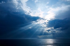 Sun through dark clouds illuminating sea. Back lit. Toned blue, monochrome